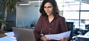 Young business woman working on laptop computer reading financial document report in office.