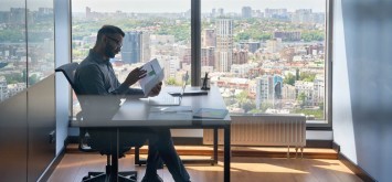 Financial analyst sitting at desk with papers working with big data, stock exchange trading operations using laptop near panoramic window in modern corporate office