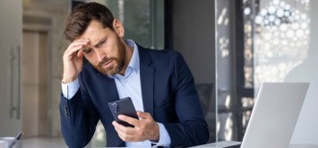 Shocked young man businessman, lawyer, banker sitting in office at desk, holding hand to head and looking frustrated at phone screen.