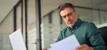 Busy middle aged executive holding documents using laptop computer in office.